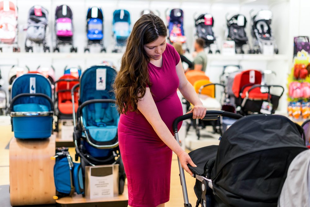 a woman in a pink dress pushing a stroller امرئه حامل تقف في قسم مستلزمات الاطفال واسكه عربة اطفال